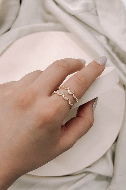 A woman's hand displays a gold double-layered ring adorned with petaled stones on a white pillow, emphasizing its adjustable size.