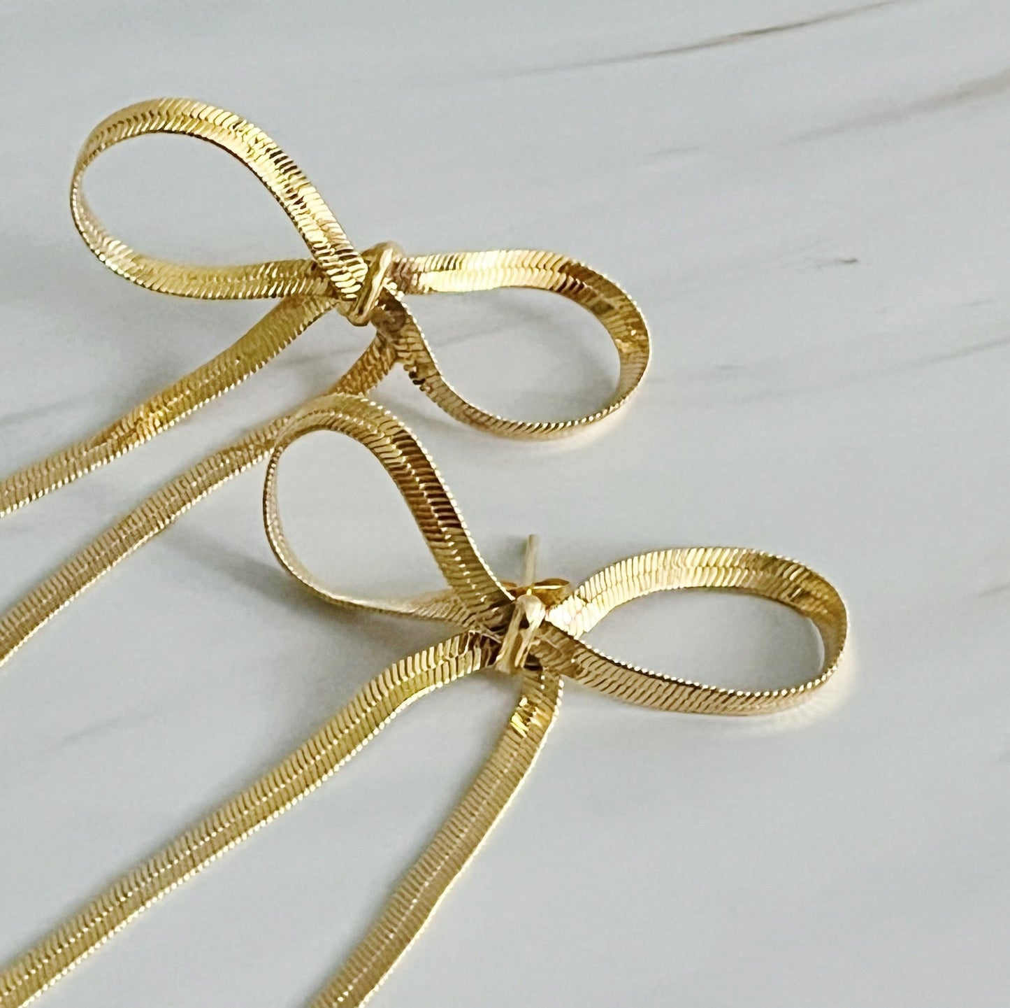 Two gold bows displayed on a white background, representing stylish long drop earrings that add sophistication to any outfit.