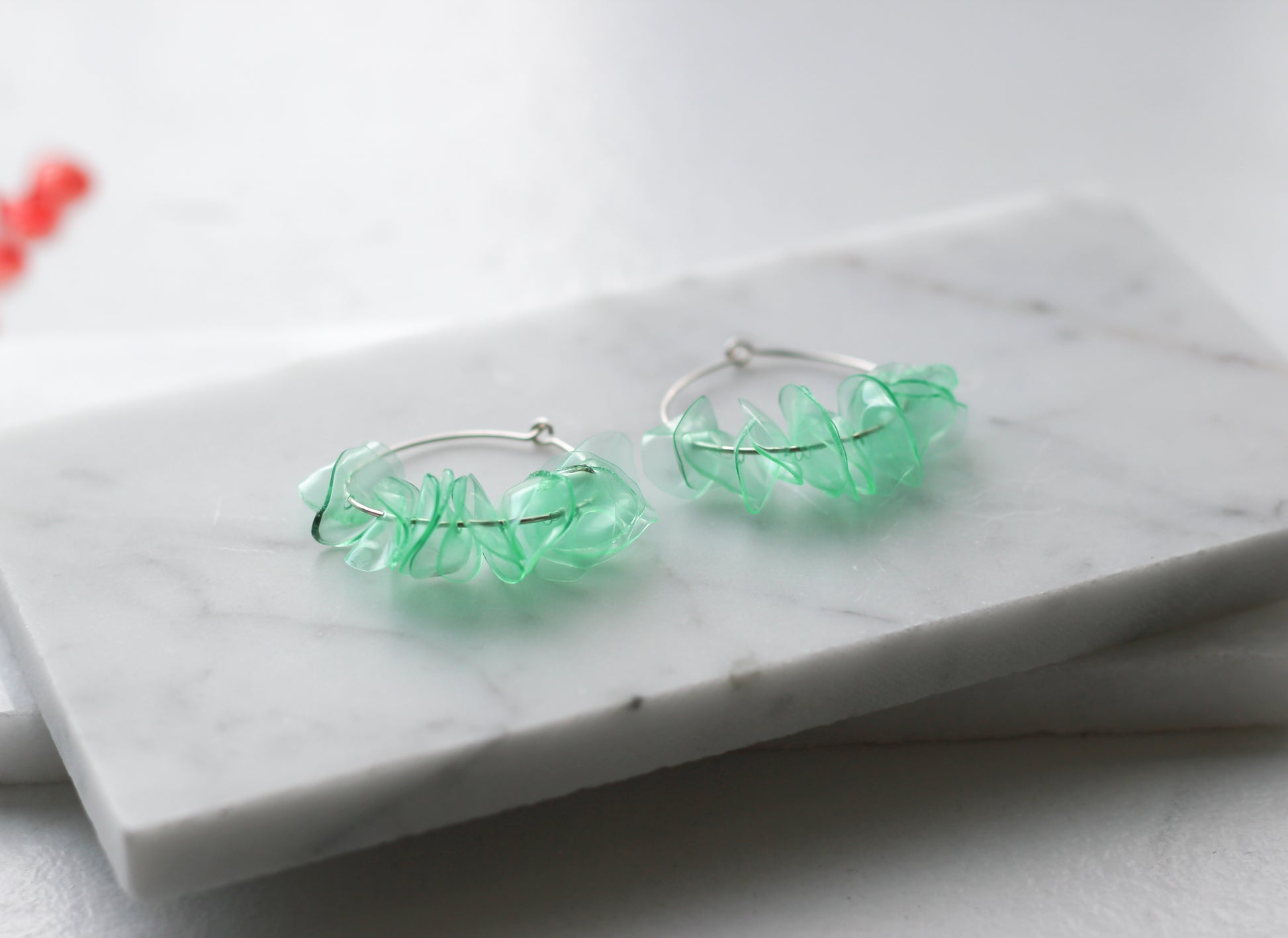  A pair of green glass earrings featuring aquamarine petals on silver hoops, resting on a marble surface. Handmade and eco-friendly.