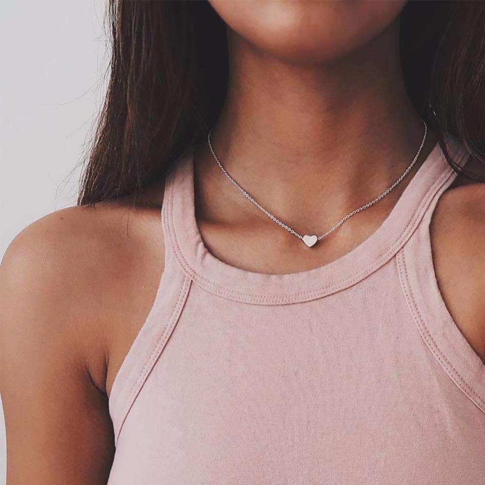 A woman in a pink tank top wears a heart pendant necklace, available in silver and gold, with a 14" length and 4" extender.