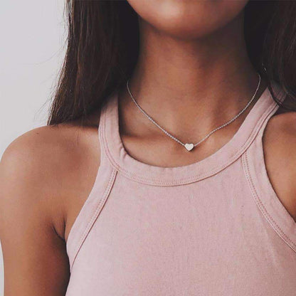 A woman in a pink tank top wears a heart pendant necklace, available in silver and gold, with a 14" length and 4" extender.