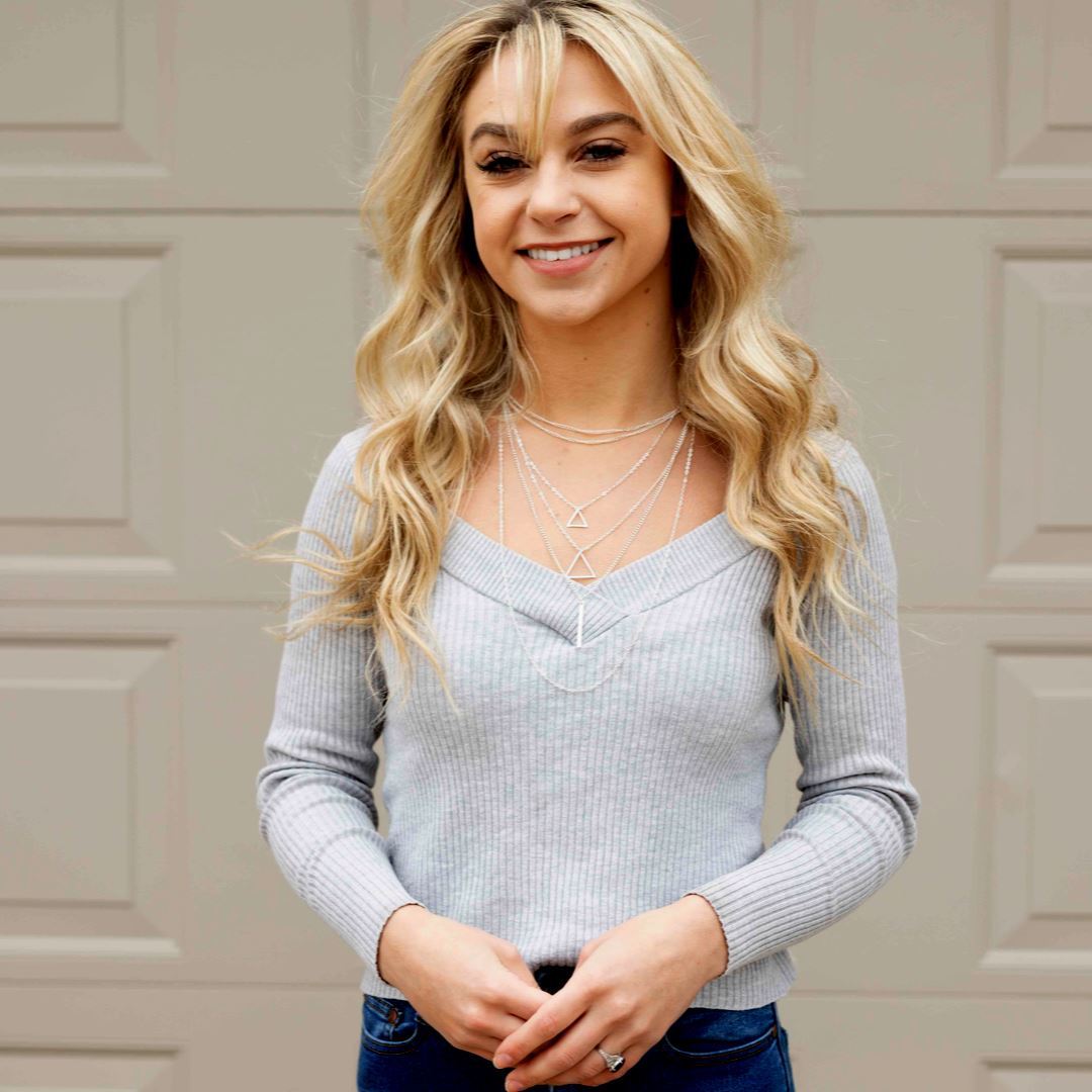  A blonde woman in jeans and a gray sweater, displaying a stylish necklace with multiple layers and adjustable chain.