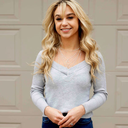  A blonde woman in jeans and a gray sweater, displaying a stylish necklace with multiple layers and adjustable chain.