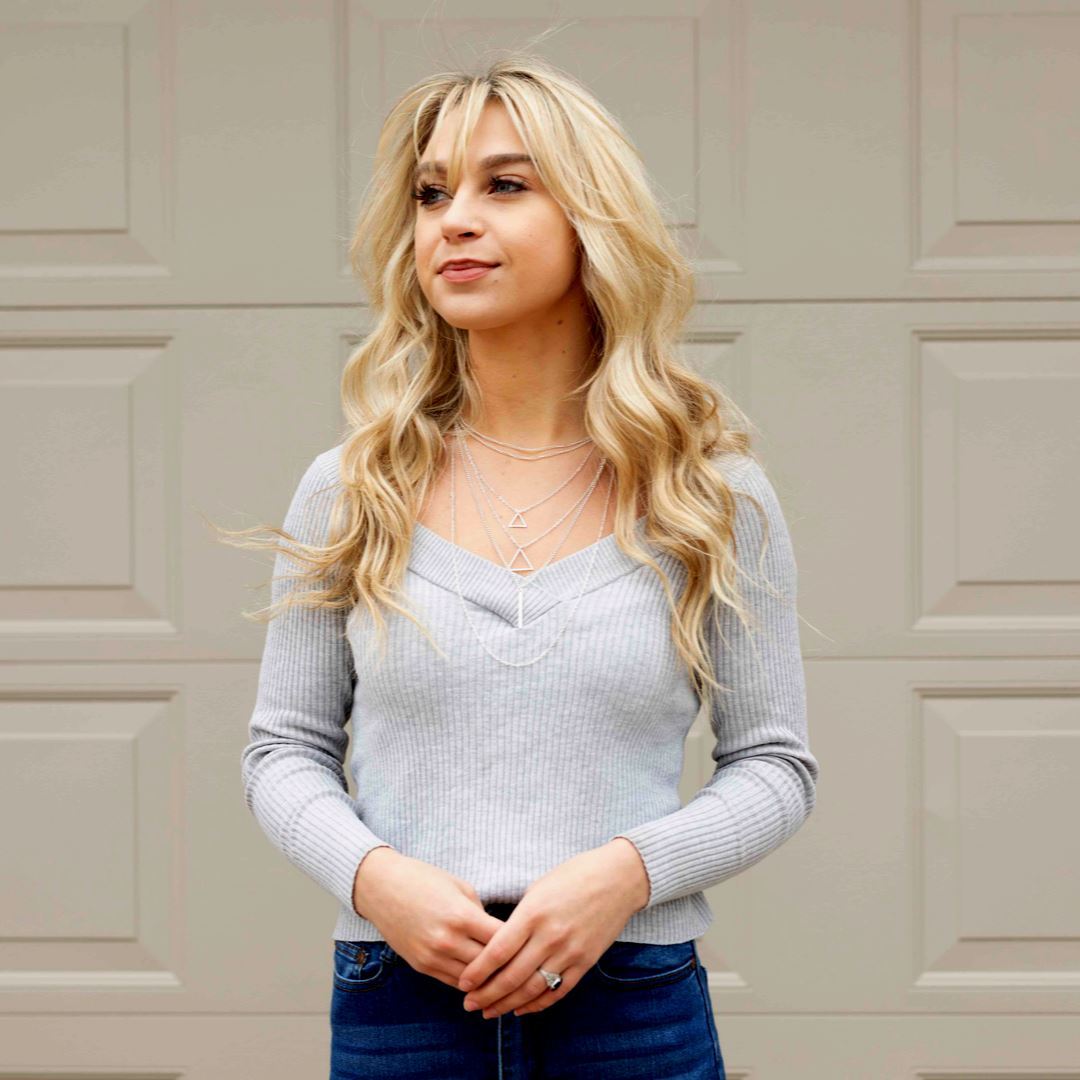 A blonde woman in jeans and a sweater showcases layered jewelry, including triangles and chokers, in front of a garage doo