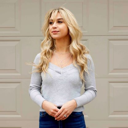 A blonde woman in jeans and a sweater showcases layered jewelry, including triangles and chokers, in front of a garage doo